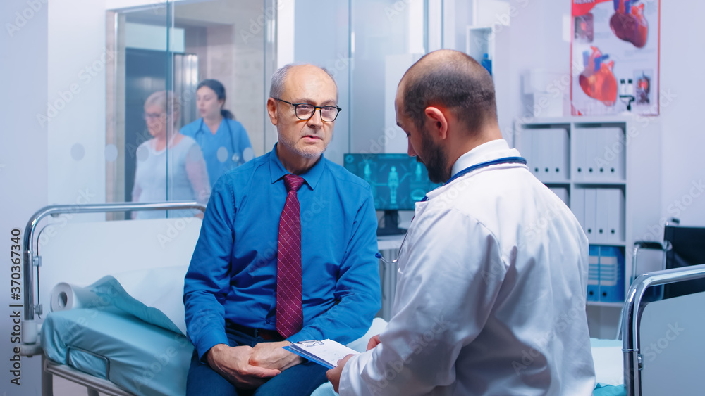 Obraz premium Senior man consulting with doctor sitting on hospital bed. Ill aged patient seeking medical advice for disease prevention from general practitioner in modern private clinic, doctor diagnosis