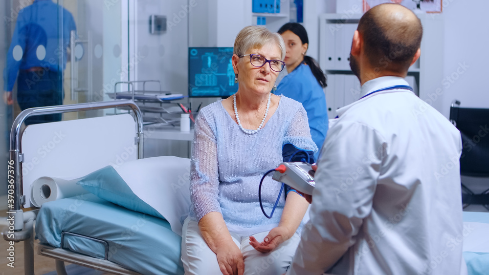 Obraz premium Practitioner measures senior retired woman blood pressure with a monitor while nurse in working in the background. Healthcare medical medicine system, treatment and diagnosis of illness examination