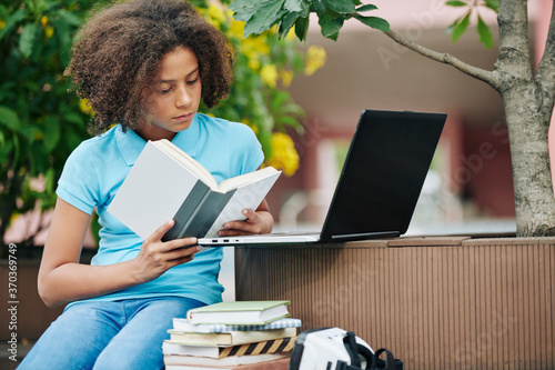 Concentrated curly mixed-race schoolgirl reading intersting book on campus photo