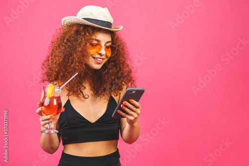 Portrait of happy woman in sunglasses using smartphone and holding cocktail isolated on pink background.