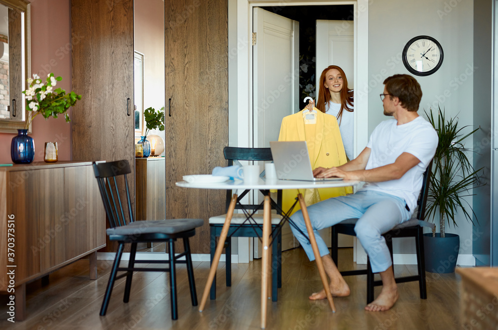 attractive lady show new clothes to working husband, young redhead woman came from shopping, share happiness with man sitting with laptop attractive lady show new clothes to working husband, young redhead woman came from shopping, share happiness with man sitting with laptop