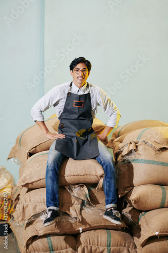 Happy proud Vietnamese worker in apron sitting on sacks with coffee her roasted