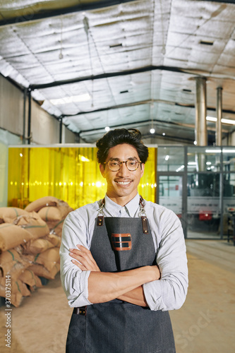Portrait of smiling confident Asian coffee roastery owner standing warehosue with sacks of coffee behend and looking at camera photo