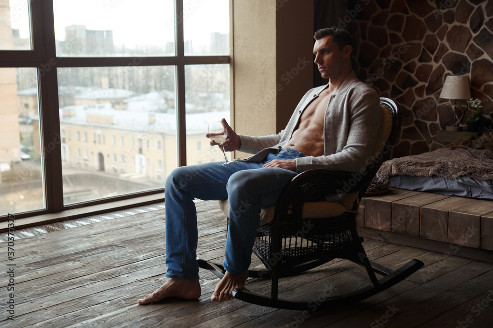 A handsome athletic man with a glass of wine in his hand is resting in a rocking chair in the interior.