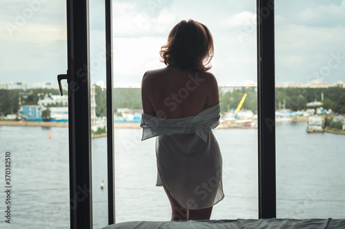 Back view of beautiful young woman in a transparent nightgown standing and enjoying near open panoramic window of apartments.