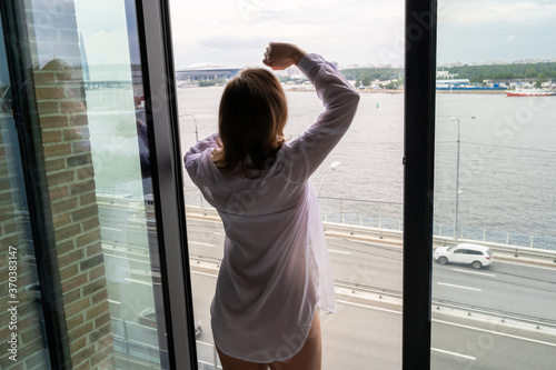 Back view of beautiful young happy woman in a transparent nightgown standing and enjoying near open panoramic window of skyscraper apartments. Female arms up over town and road. © flowertiare