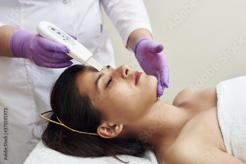 Woman receiving cleansing therapy with a professional ultrasonic equipment in cosmetology office. Procedure of ultrasonic cleaning of face. Cosmetological clinic
