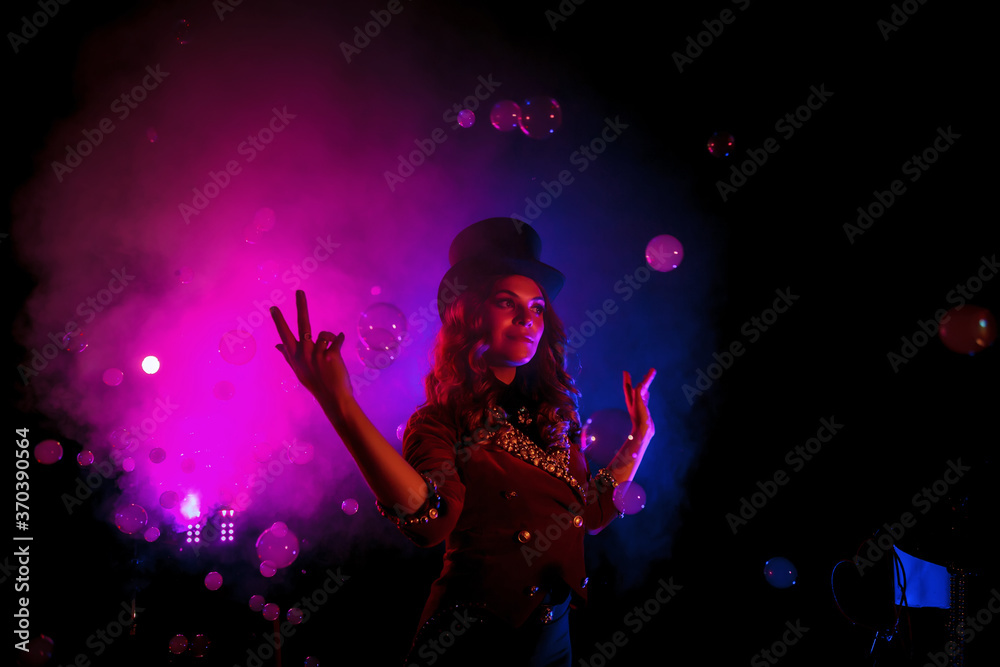 Female magician, an illusionist in theatrical clothes, makes show with soap bubbles on black background. Girl actress in stage costume and top hat on her head. Concept of theatrical performance