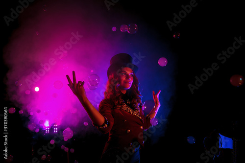 Female magician, an illusionist in theatrical clothes, makes show with soap bubbles on black background. Girl actress in stage costume and top hat on her head. Concept of theatrical performance