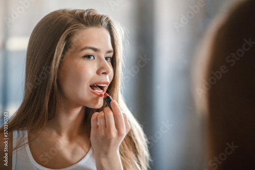 Attractive young lady applying lipstick on her lips near mirror at home, blank space