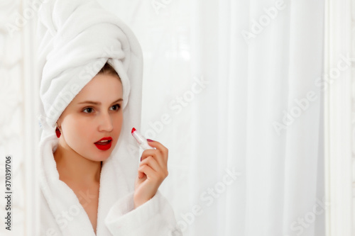 Woman paints her lips in hotel bathroom after shower. Pretty female of Slavic appearance in dressing gown about mirror. Happy lady. Healthy facial skin. Cute girl washed her face and shows emotions