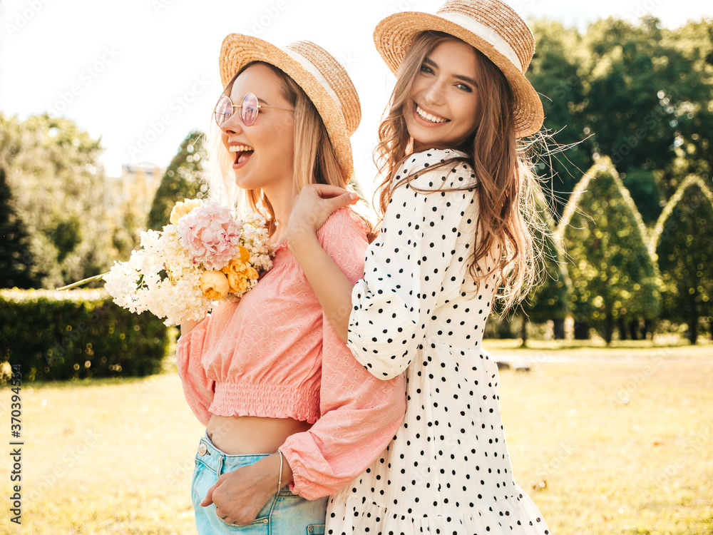 Two young beautiful smiling hipster woman in trendy summer sundress. Sexy carefree women posing in the park in hats. Positive models having fun and hugging at sunset. They holding flowers