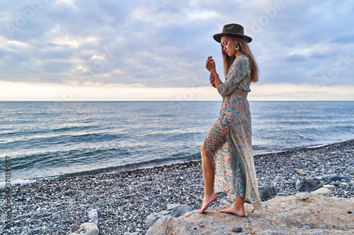 Boho woman in long dress and felt hat standing on stones by the sea