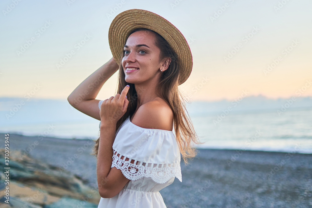 Portrait of beautiful young happy smiling romantic woman in a summer white dress with bare shoulders and a straw hat by the seashore at summertime Portrait of beautiful young happy smiling romantic woman in a summer white dress with bare shoulders and a straw hat by the seashore at summertime