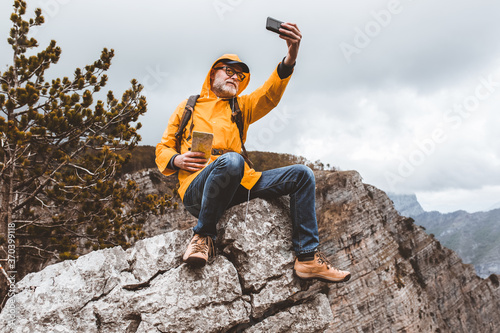 Middle age man traveler in raincoat and backpack enjoying view of mountains make selfie photo.