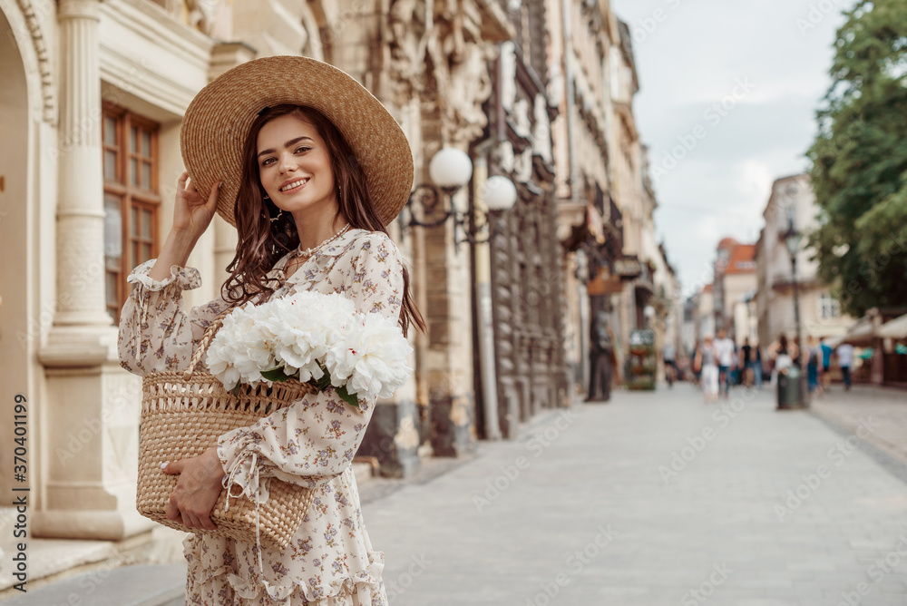 Happy smiling woman wearing straw hat, floral print dress, holding wicker bag with white peony flowers, posing in street of European city. Lifestyle, travel conception. Copy, empty space for text