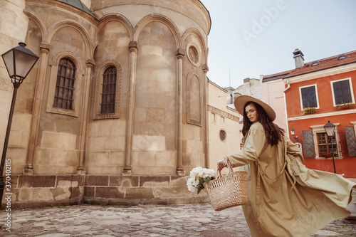 Canvas Print Beautiful woman wearing straw hat, long trench coat, holding wicker bag with flowers, walking in street of European city