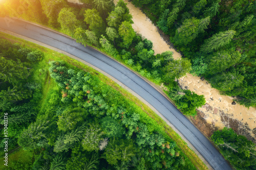 Aerial view of road in beautiful green forest at sunset in summer. Colorful landscape with roadway, pine trees, blurred car and river in Carpatian mountains. Top view of highway. Travel in Ukraine