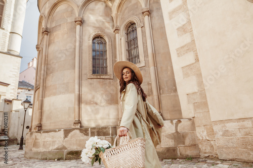 Young elegant lady wearing straw hat, long trench coat, holding wicker bag with flowers, walking in street of European city. Lifestyle, travel conception. Copy, empty space for text