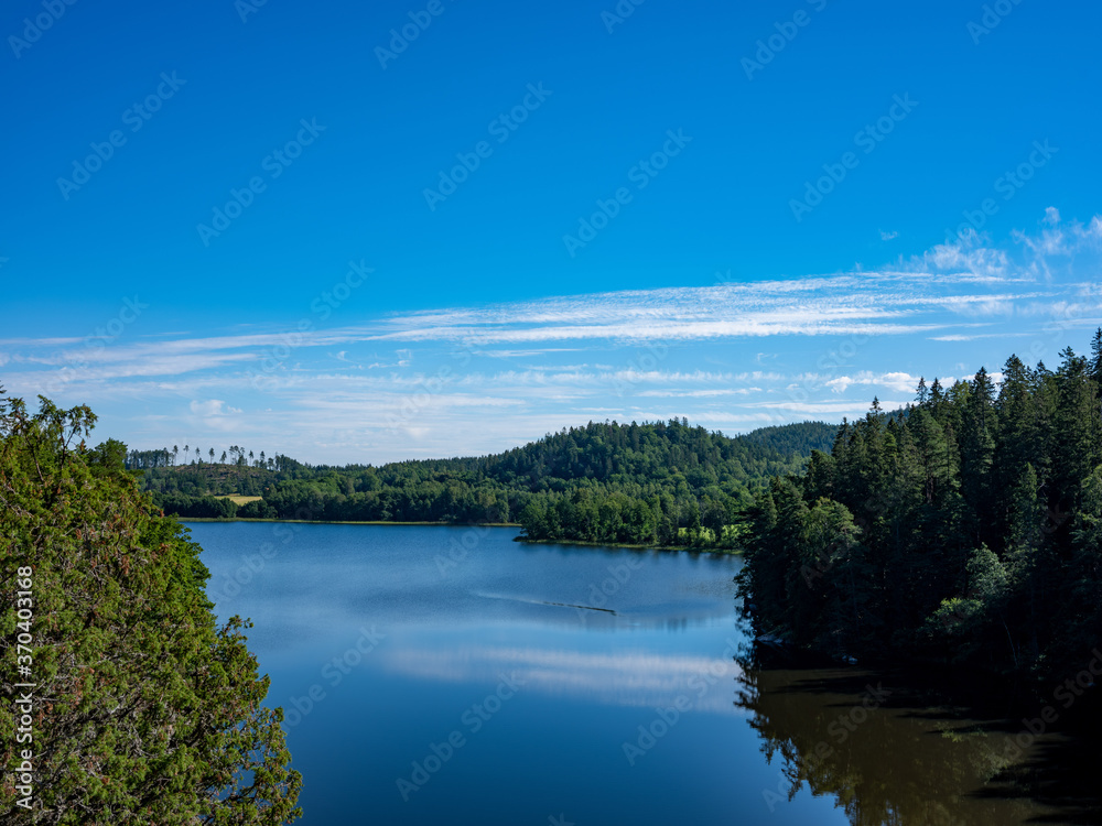 Scenic landscape photo of a blue lake surrounded by lush forest. It is a sunny summers day with blue sky. The water is calm.