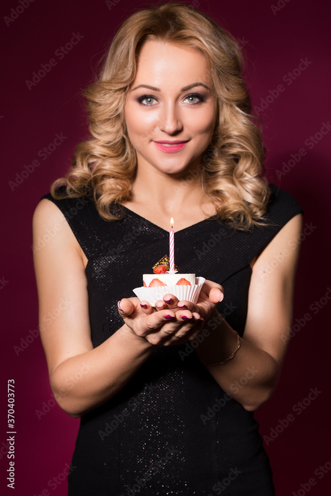 Beautiful blonde woman in luxury black dress and curly hairstyle blowing candle on birthday cake. clear skin. dark red background Beautiful blonde woman in luxury black dress and curly hairstyle blowing candle on birthday cake. clear skin. dark red background