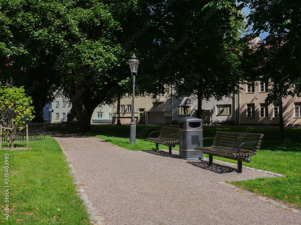 View of a pathway through a park.
Two benches, a waste basket and an old lamp post is placed beside the pathway.