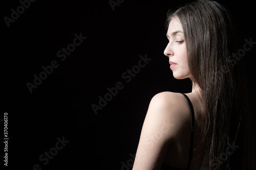 A long-haired girl on a dark background. A girl in an open dress stands with her back to the camera. Serious woman on a black background. The girl looks down.