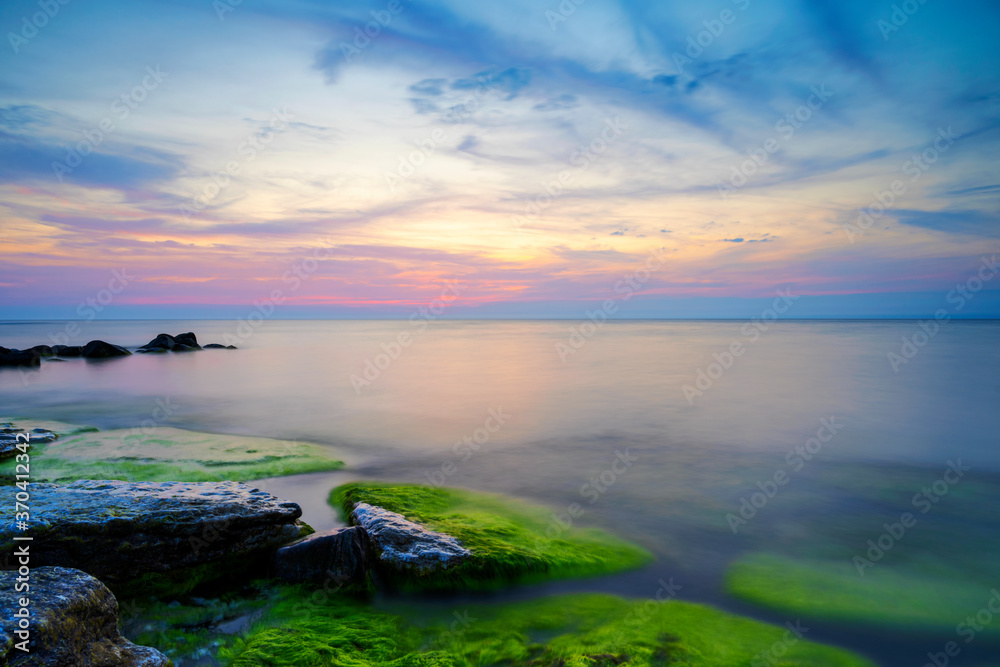 Naklejka premium Cloudy summer sunset reflecting in ocean with endless horizon and deep blue ocean, silhouette of boulders laying in the foreground in shallow water at island of Gotland, Sweden