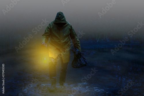 A scary man with wading boots , rain jacket and a bag wades by candlelight through the foggy swamp..