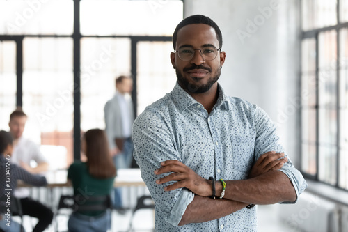 Head shot portrait confident young African American businessman wearing glasses looking at camera, successful executive startup founder standing in modern office room with arms crossed photo