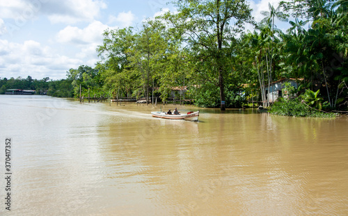 Duas pessoas andando de barco de madeira a motor em rio na margem da floresta amazônica, no Pará. Casas ribeirinhas na margem da água.
