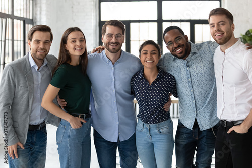 Smiling diverse business people, successful team, staff members hugging, standing in modern office, looking at camera, happy overjoyed employees colleagues posing for corporate photo