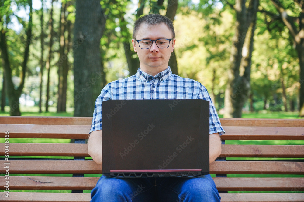 Fototapeta premium The guy sits on a park bench and works at a laptop.
