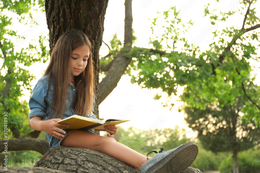 Cute little girl reading book on tree in park - obrazy, fototapety, plakaty 