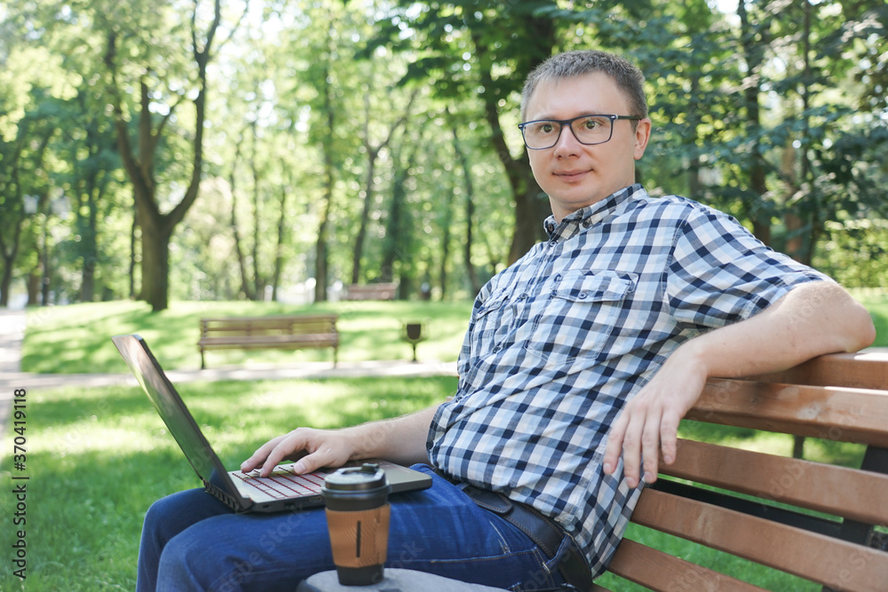 Fototapeta premium A guy with a laptop sits on a park bench. A cup of coffee stands near the laptop.