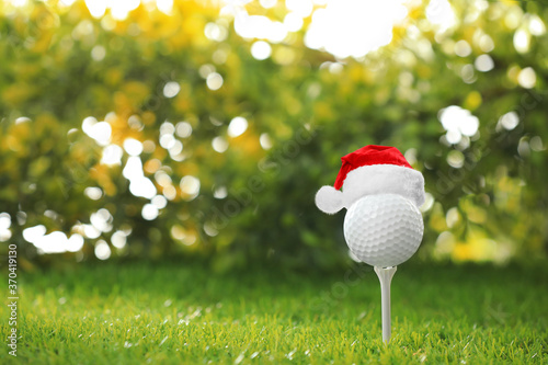 Golf ball with small Santa hat on tee against blurred background. Space for text
