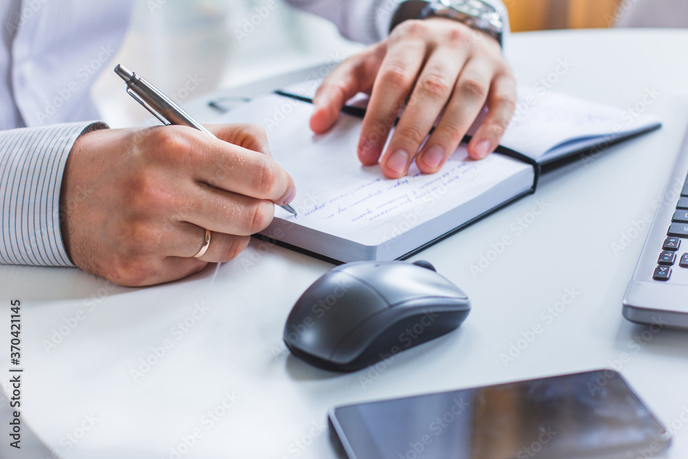 An image of the hands of a man sitting at a table and writing in a Notepad with a ballpoint pen. Next to it is a laptop, a computer mouse, and a cell phone An image of the hands of a man sitting at a table and writing in a Notepad with a ballpoint pen. Next to it is a laptop, a computer mouse, and a cell phone