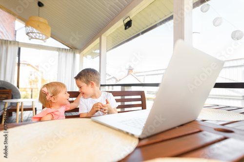 Small smiling kids brother and sister laugh and play laptop, communicate video conference chatting. Spacious cozy home interior.