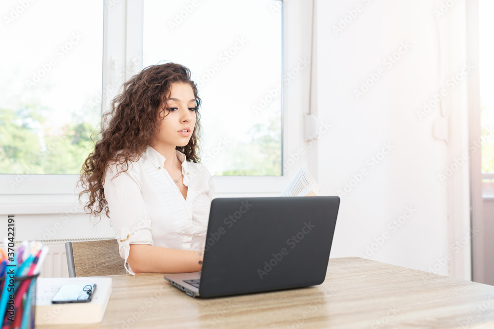 Successful female with long brown hair in white shirt sitting in office by the window and looking at laptop. Successful female with long brown hair in white shirt sitting in office by the window and looking at laptop.