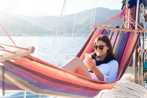 Woman relaxing on yacht in beach hammock. Traveler with mobile phone. Travel on sailboat. Enjoying summer vacation. Freelance safe workplace  distance learning. Successful business lifestyle.