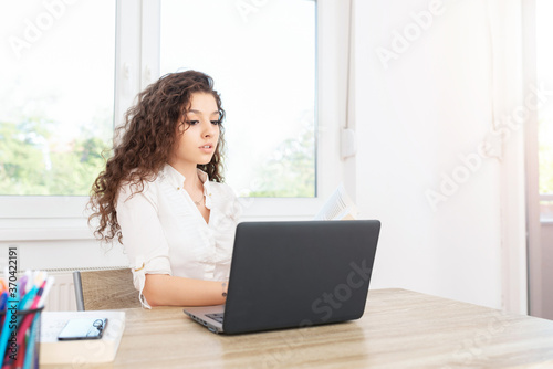 Successful female with long brown hair in white shirt sitting in office by the window and looking at laptop.