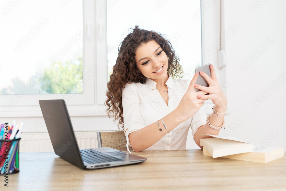 Attractive lady in white shirt sitting at work desk with laptop, looking at cellphone and smiling. Attractive lady in white shirt sitting at work desk with laptop, looking at cellphone and smiling.