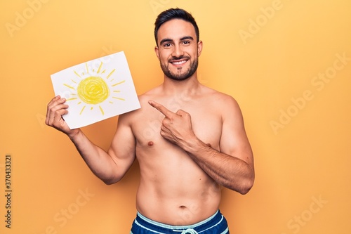 Young handsome man with beard on vacation wearing swimwear holding paper with sun draw smiling happy pointing with hand and finger