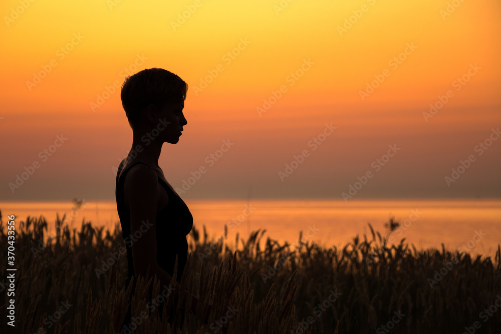 silhouette portrait of a woman standing in the grass, in profile, headshot, in the rays of the setting sun over the sea, the concept of tranquility, relaxation and unity with nature