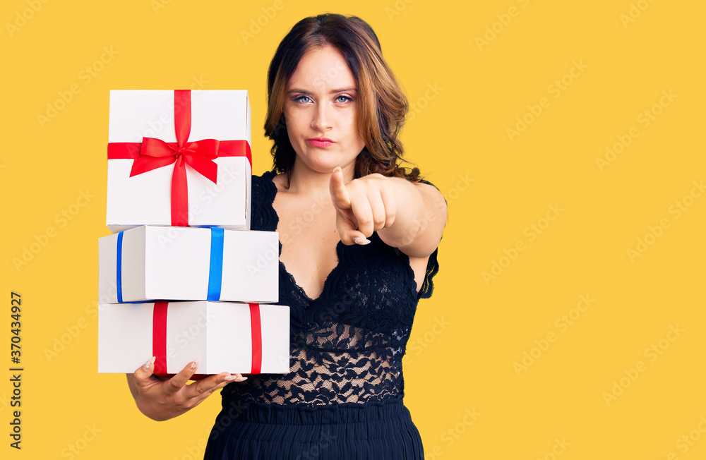 Young beautiful caucasian woman holding gifts pointing with finger to the camera and to you, confident gesture looking serious Young beautiful caucasian woman holding gifts pointing with finger to the camera and to you, confident gesture looking serious