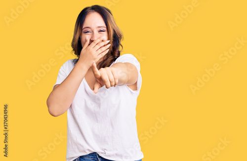 Young beautiful caucasian woman wearing casual white tshirt laughing at you, pointing finger to the camera with hand over mouth, shame expression