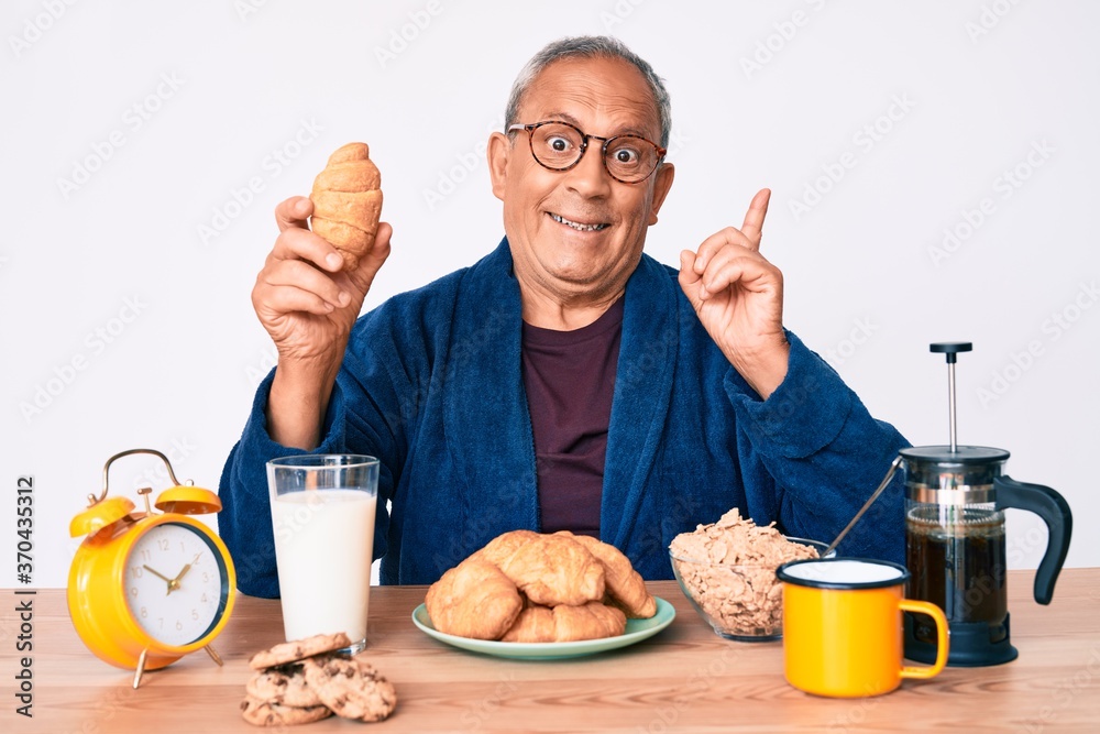 Senior handsome man with gray hair sitting on the table eating croissant for breakfast surprised with an idea or question pointing finger with happy face, number one Senior handsome man with gray hair sitting on the table eating croissant for breakfast surprised with an idea or question pointing finger with happy face, number one