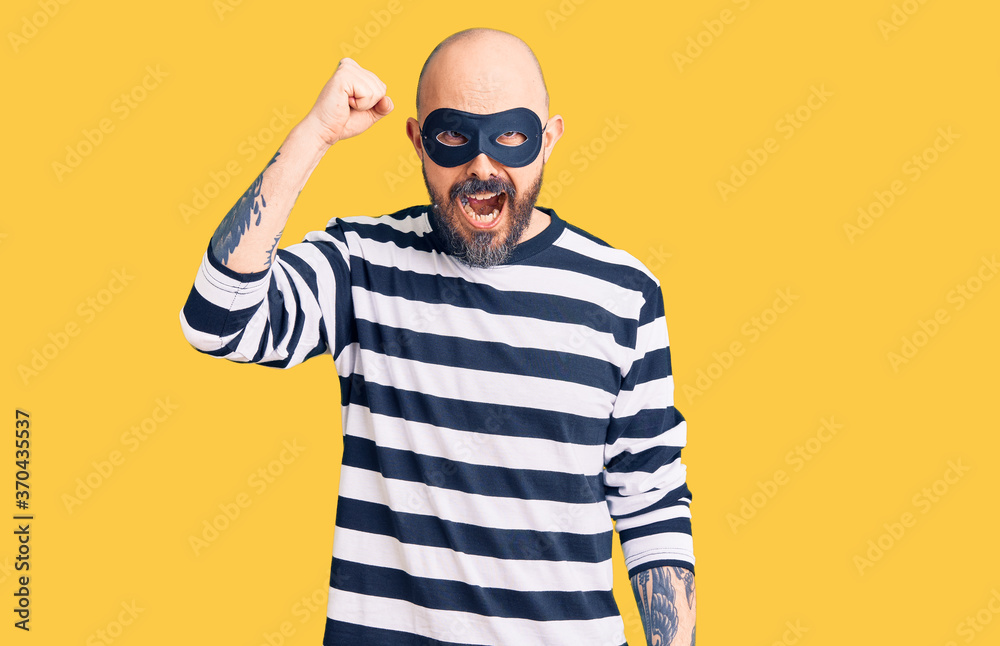 Young handsome man wearing burglar mask angry and mad raising fist frustrated and furious while shouting with anger. rage and aggressive concept. Young handsome man wearing burglar mask angry and mad raising fist frustrated and furious while shouting with anger. rage and aggressive concept.