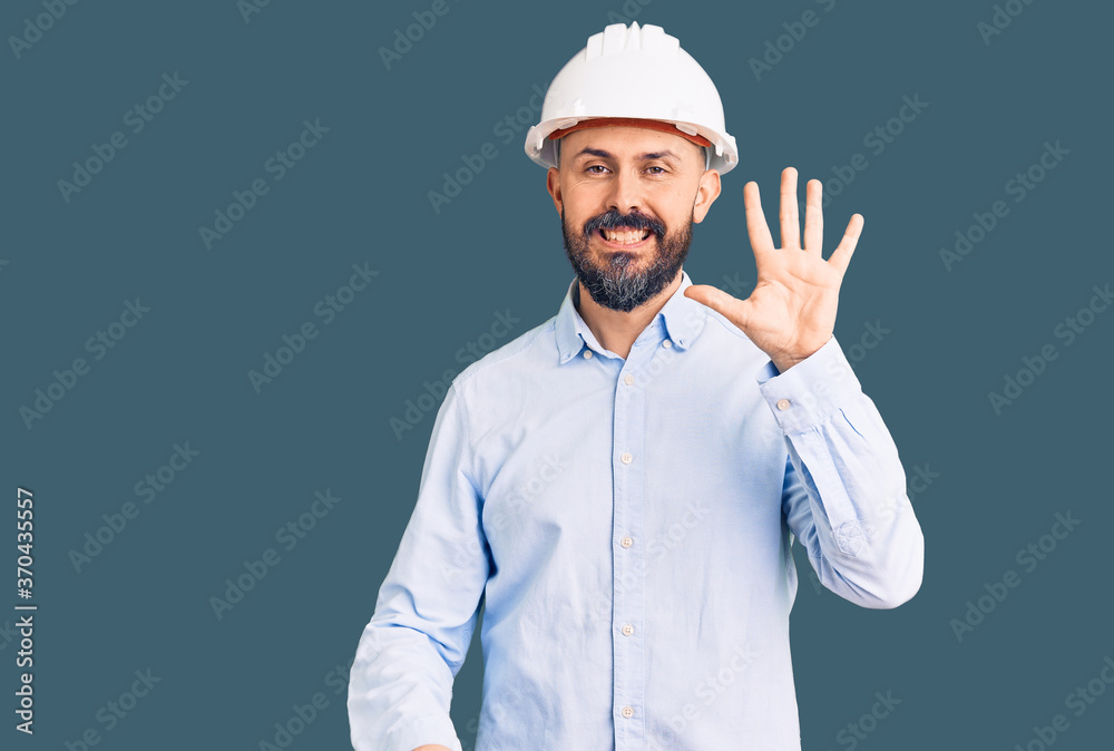 Young handsome man wearing architect hardhat showing and pointing up with fingers number five while smiling confident and happy. Young handsome man wearing architect hardhat showing and pointing up with fingers number five while smiling confident and happy.