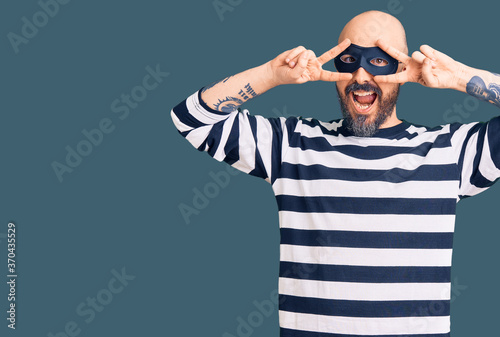 Young handsome man wearing burglar mask doing peace symbol with fingers over face, smiling cheerful showing victory © Krakenimages.com